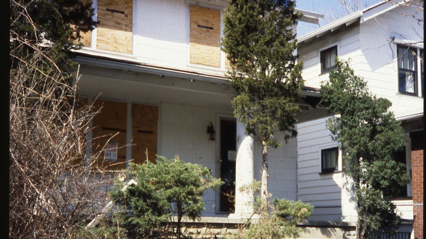 Vacant white house with plywood on the windows and an overgrown front yard
