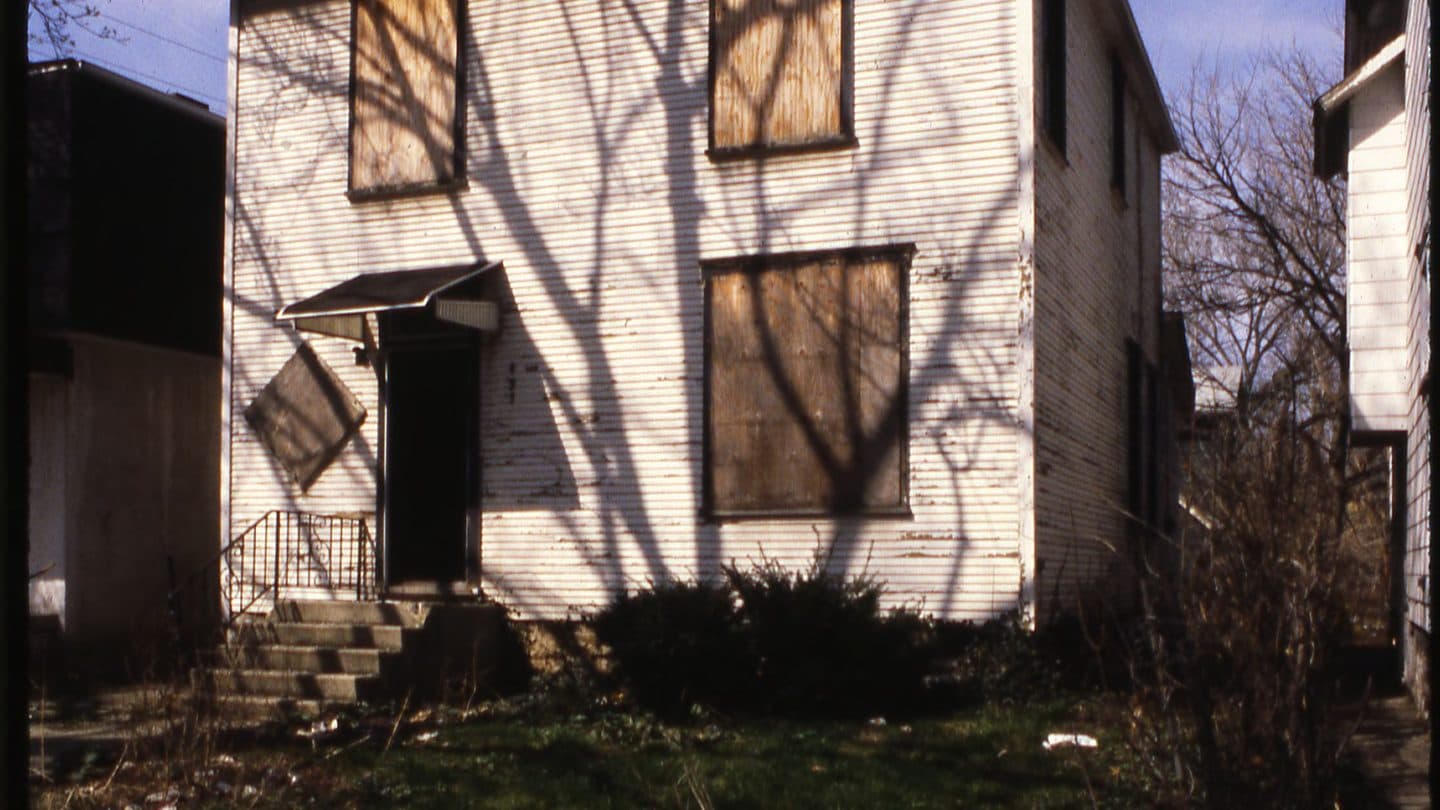 Vacant two-story house with plywood on the windows and litter in the yard