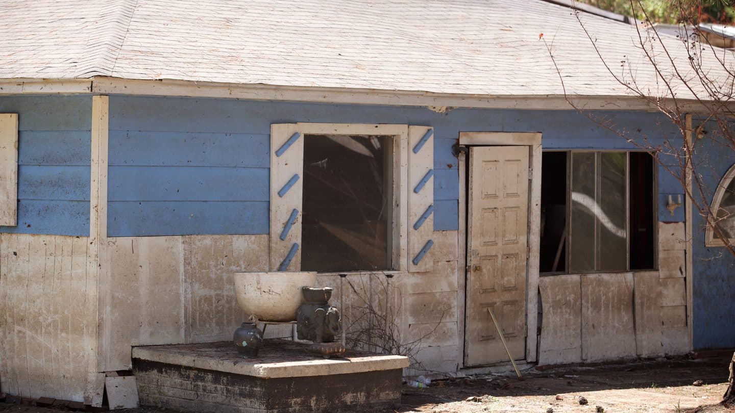 Neglected one-story house with stained siding, an open door, and debris in the yard