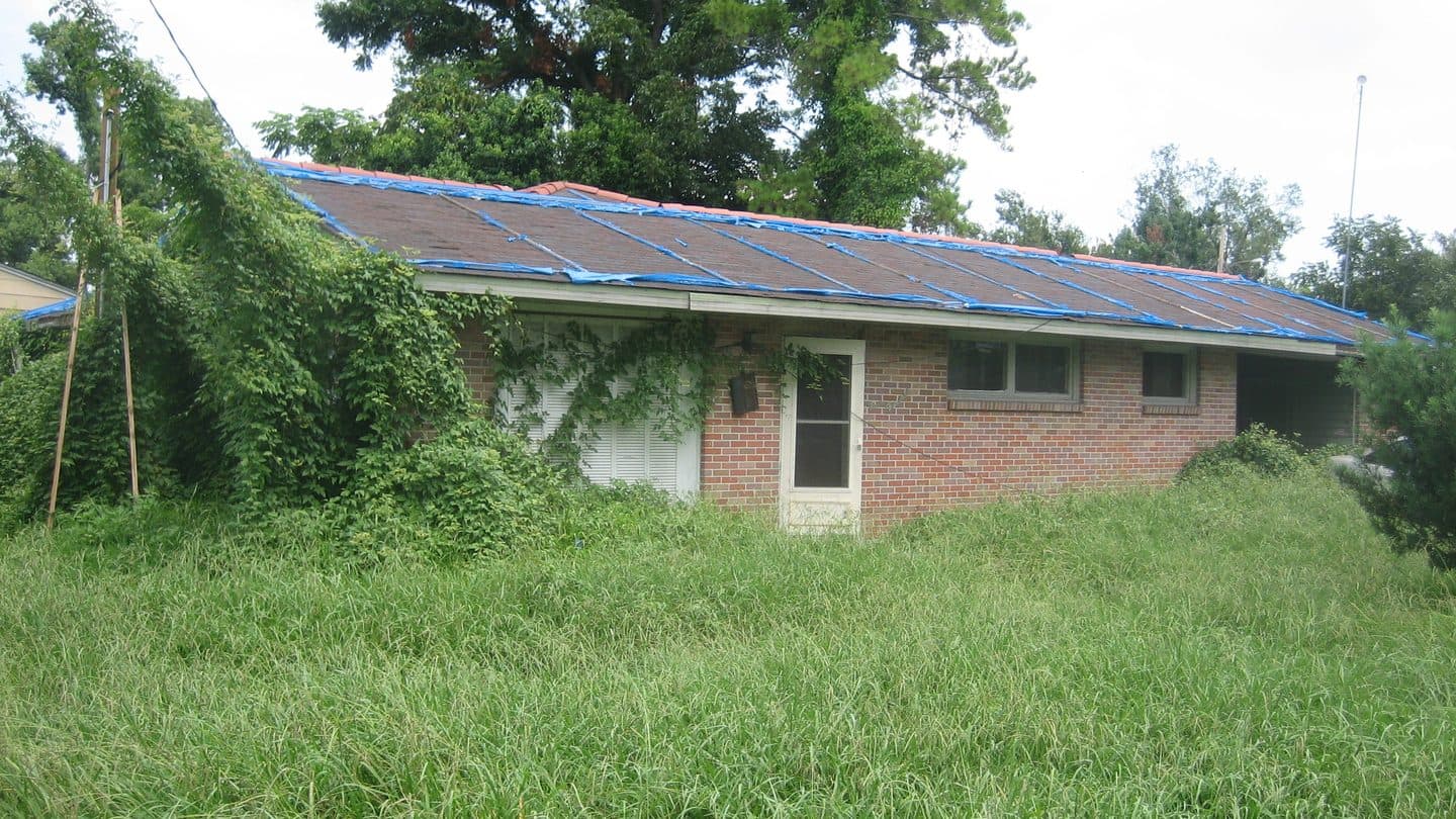 Brick house with blue tarps covering a failed roof and an overgrown yard
